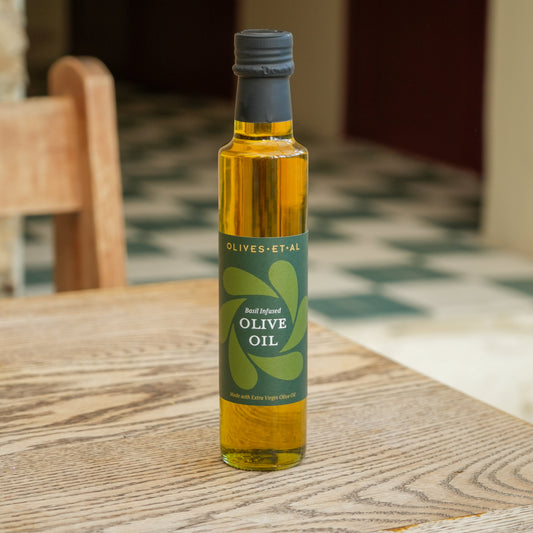 A bottle of OLIVES-ET-AL Basil olive oil standing on a wooden table in a home setting with a blurred checkered floor and chair in the background.