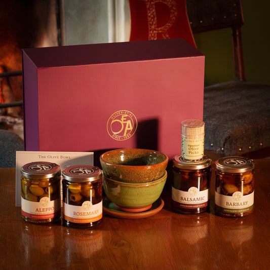 Olives Et Al Table Hamper presented in a purple gift box, featuring a green hand-thrown Dorset Olive Bowl, four jars of marinated olives and Signature Olive Picks. The selection includes aromatic Barbary Olives, gently spicy Aleppo Olives, Balsamic Olives and fragrant Rosemary Olives. The ceramic bowl separates olives from their extra virgin olive oil, ready for dipping bread or dressing salads. A thoughtful Christmas gift for olive lovers, perfect for festive snacks, drinks and gatherings around the table.