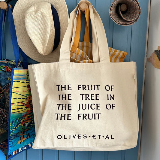 An off-white canvas tote bag with black text reading 'THE FRUIT OF THE TREE IN THE JUICE OF THE FRUIT' and 'OLIVES•ET•AL' at the bottom, hanging on a wooden peg against a blue paneled wall. A straw hat, a rolled woven mat, and other hanging bags are visible around it.