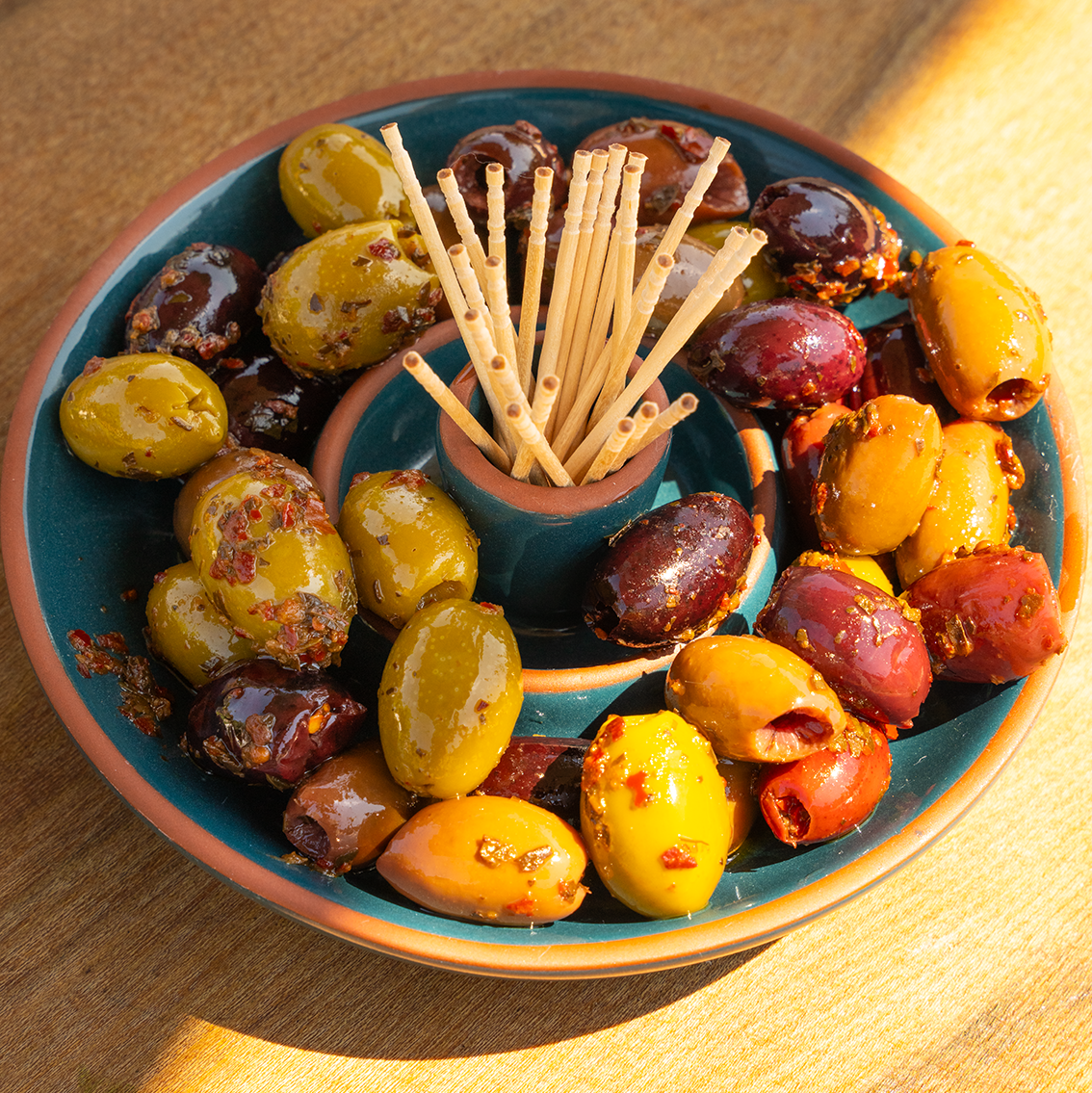 A round teal ceramic serving platter filled with assorted marinated olives in green, red, and purple, with a small center well holding wooden toothpicks, set on a sunlit wooden surface.
