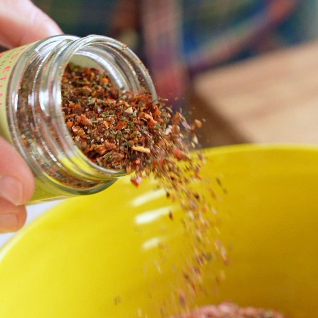 Hand pouring spices from a jar into a yellow bowl.