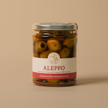 A glass jar of Aleppo olives in extra virgin olive oil, featuring a white and red label with the brand name 'Aleppo' and a silver metal lid, set against a solid beige background.