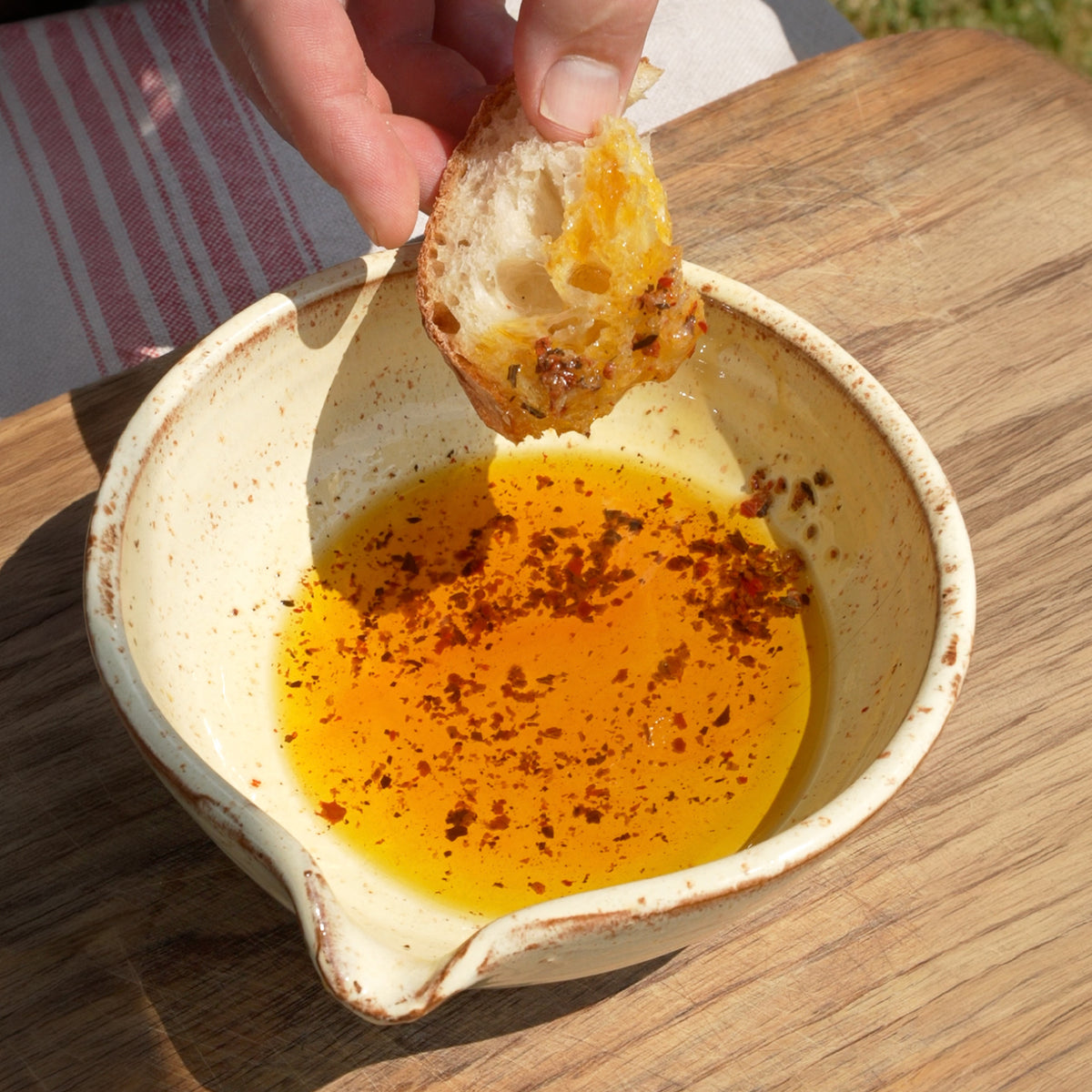 A hand holds a piece of toasted bread dipped into a small ceramic bowl of golden olive oil oil with red spice flakes, set on a wooden table outdoors.