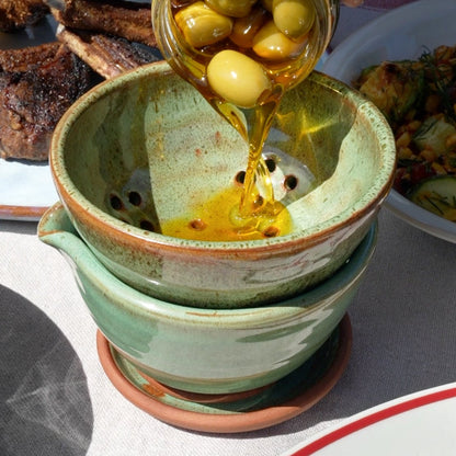 A hand pouring golden olive oil from a glass jar containing green olives into a stacked ceramic funnel and catch basin set outdoors on a table with food in the background.
