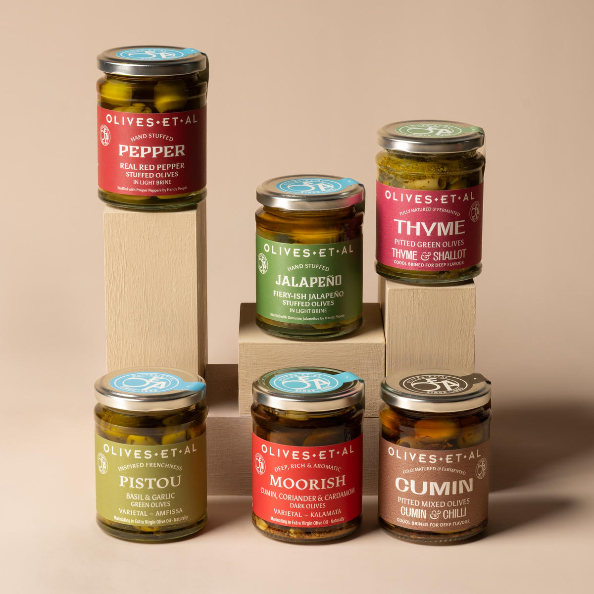 Six glass jars from the Olives Et Al Best Selling Olive Bundle. The colourful collection includes Fiery Jalapeño stuffed olives, Thyme & Shallot pitted green olives, Moorish dark olives with cumin, coriander and cardamom, Real Red Pepper stuffed olives, Cumin & Chilli pitted mixed olives, and Pistou green olives with basil and garlic.