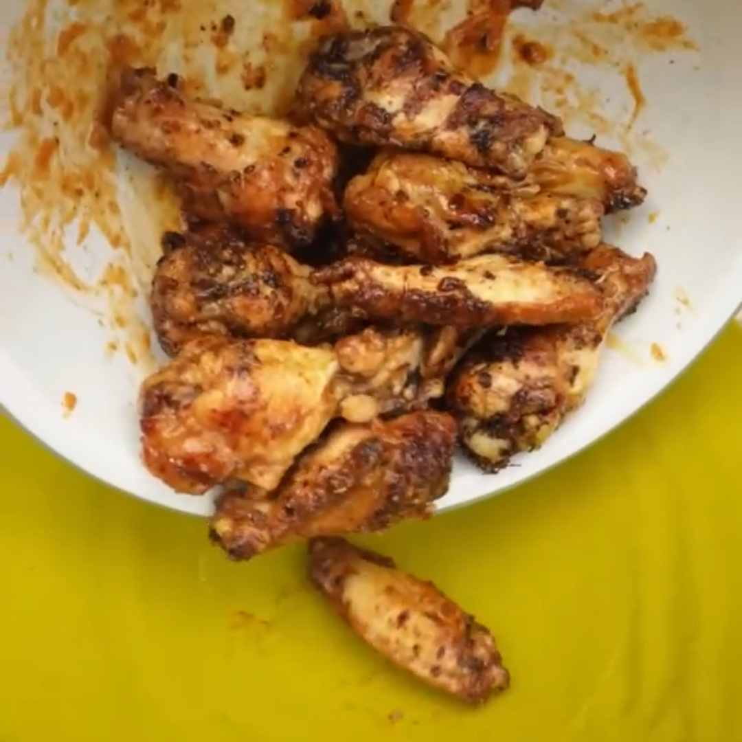 Grilled chicken wings with a reddish-brown glaze being poured from a white bowl onto a yellow plate, with a hand holding the bowl.