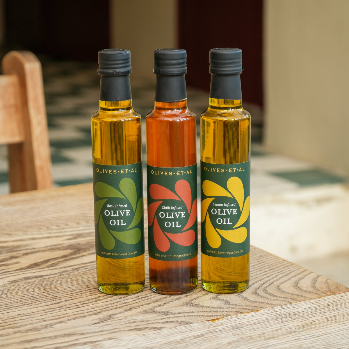 Three bottles of olive oil from Olives d'Etal standing on a wooden table. The left bottle has a green label, the middle bottle has a red label, and the right bottle has a yellow label.