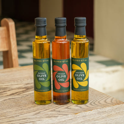 Three bottles of olive oil from Olives d'Etal standing on a wooden table. The left bottle has a green label, the middle bottle has a red label, and the right bottle has a yellow label.