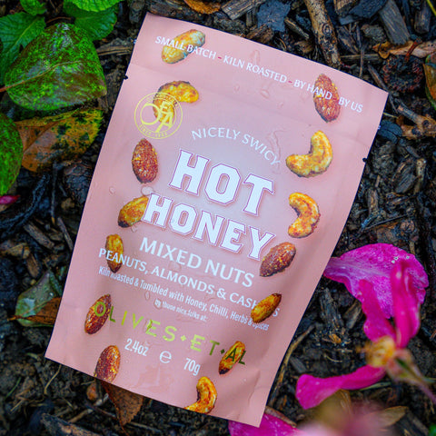 Pink package of 'Nicely Swicy Hot Honey Mixed Nuts' on a natural background with leaves and flowers.