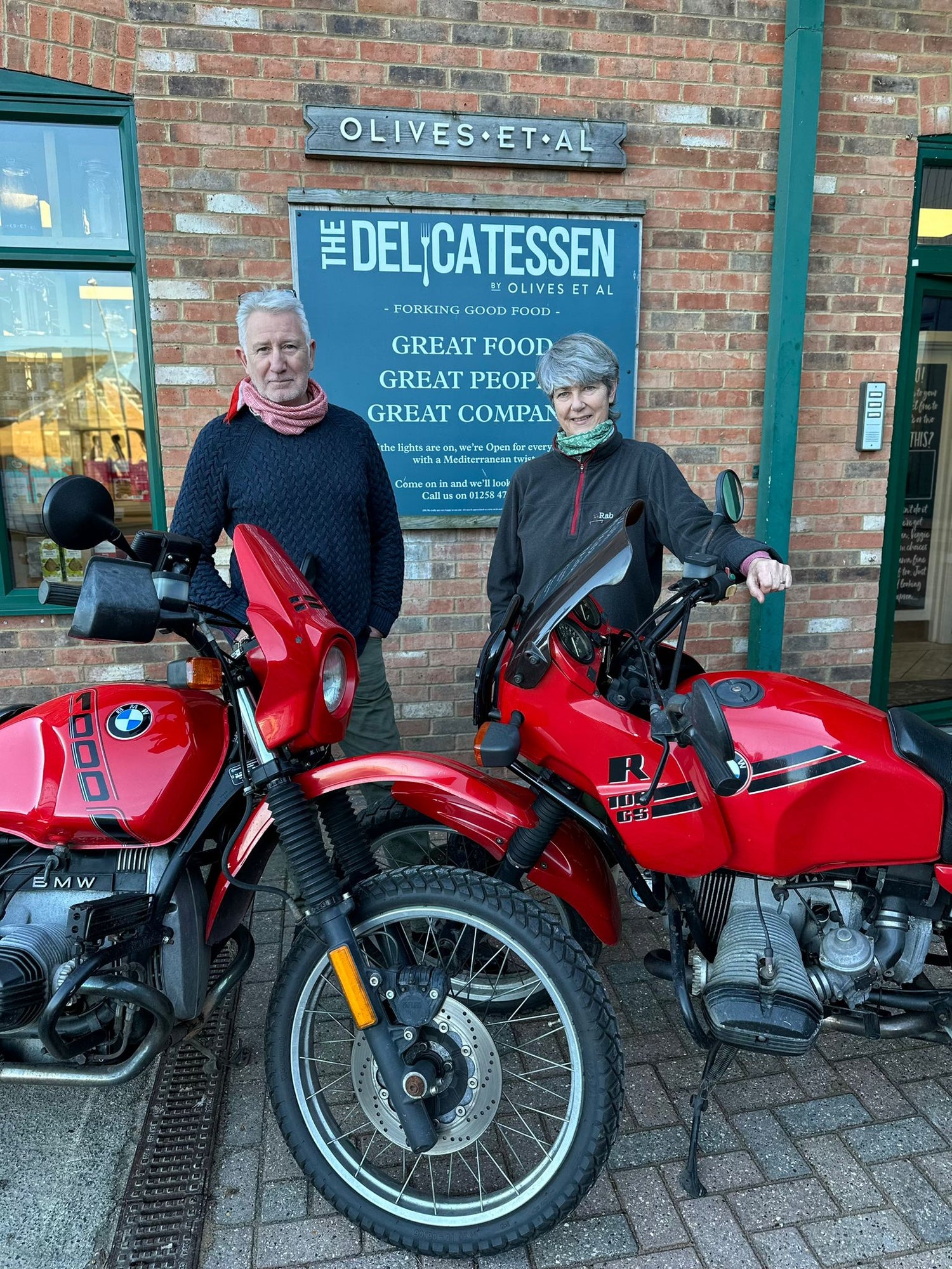 Two people standing behind two red BMW motorcycles in front of a brick storefront with a sign for The Delicatessen.