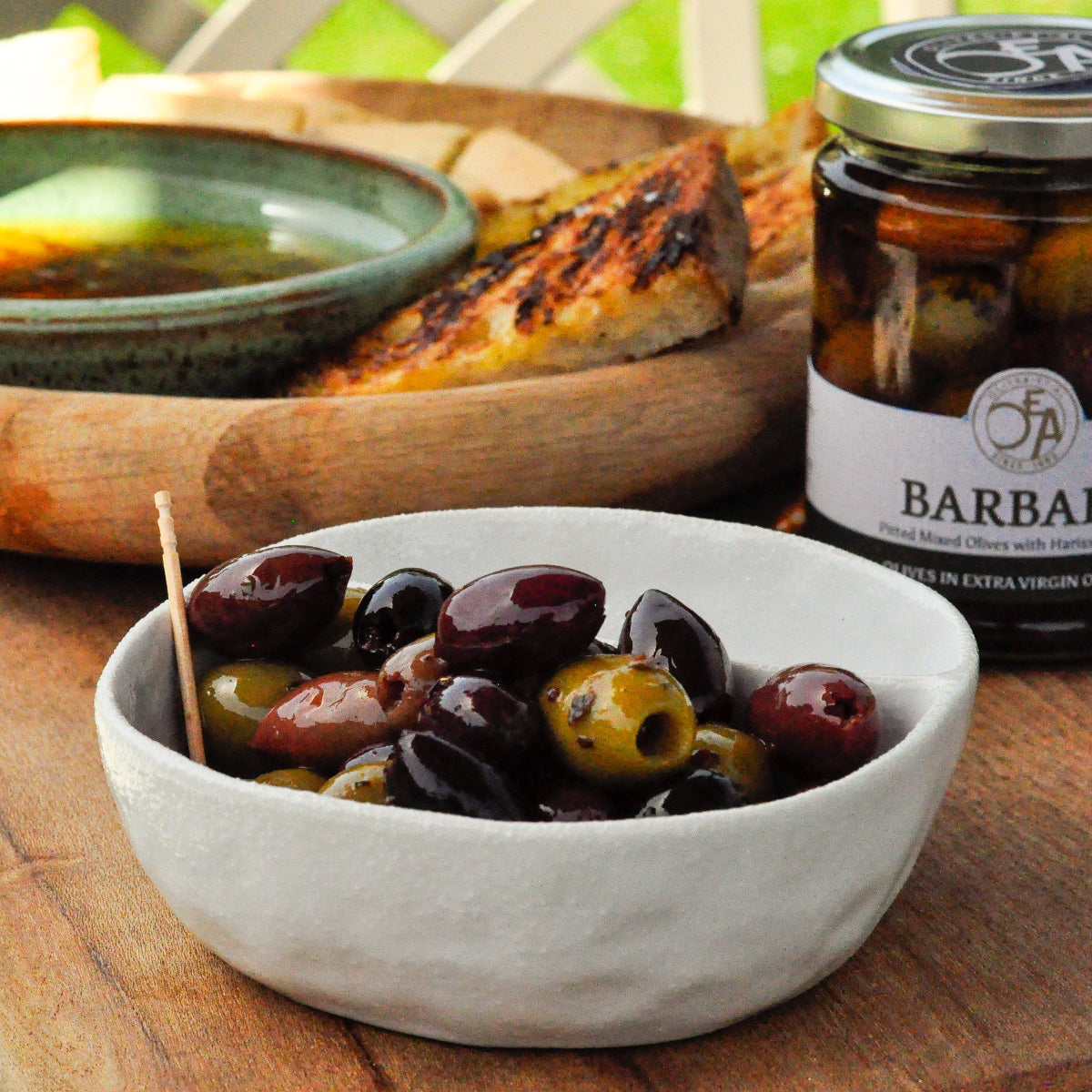 Jar of Barbary olives on a wooden table with a bowl of olives and bread.