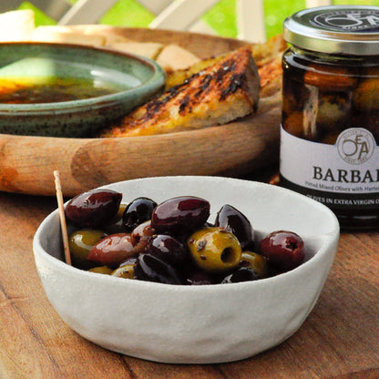 Jar of Barbary olives on a wooden table with a bowl of olives and bread.