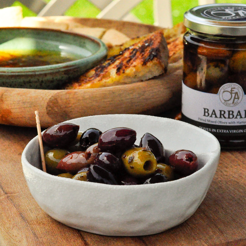 Jar of Barbary olives on a wooden table with a bowl of olives and bread.