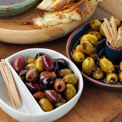 Assorted olives in a white divided bowl with bread and a bowl of snacks on a wooden table.