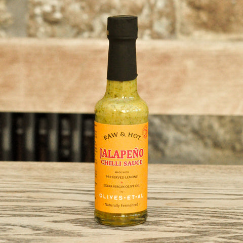 Bottle of Raw & Hot Jalapeño Chilli Sauce on a wooden surface with a stone wall background