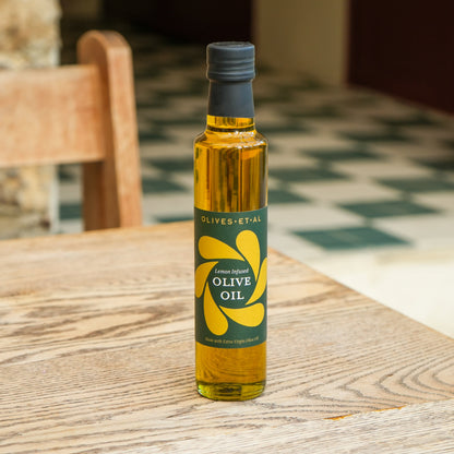 A bottle of Olives-et-al lemon infused olive oil stands on a wooden table in a warm, softly blurred indoor setting.