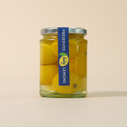 A glass jar of preserved lemons with a blue and yellow label, set against a solid beige background.