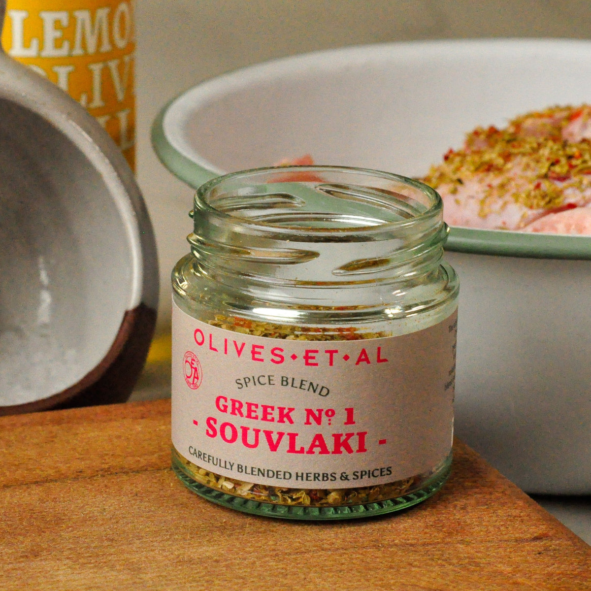 Jar of 'Greek No. 1 Souvlaki' spice blend by Olives Et Al on a wooden surface with a blurred background.