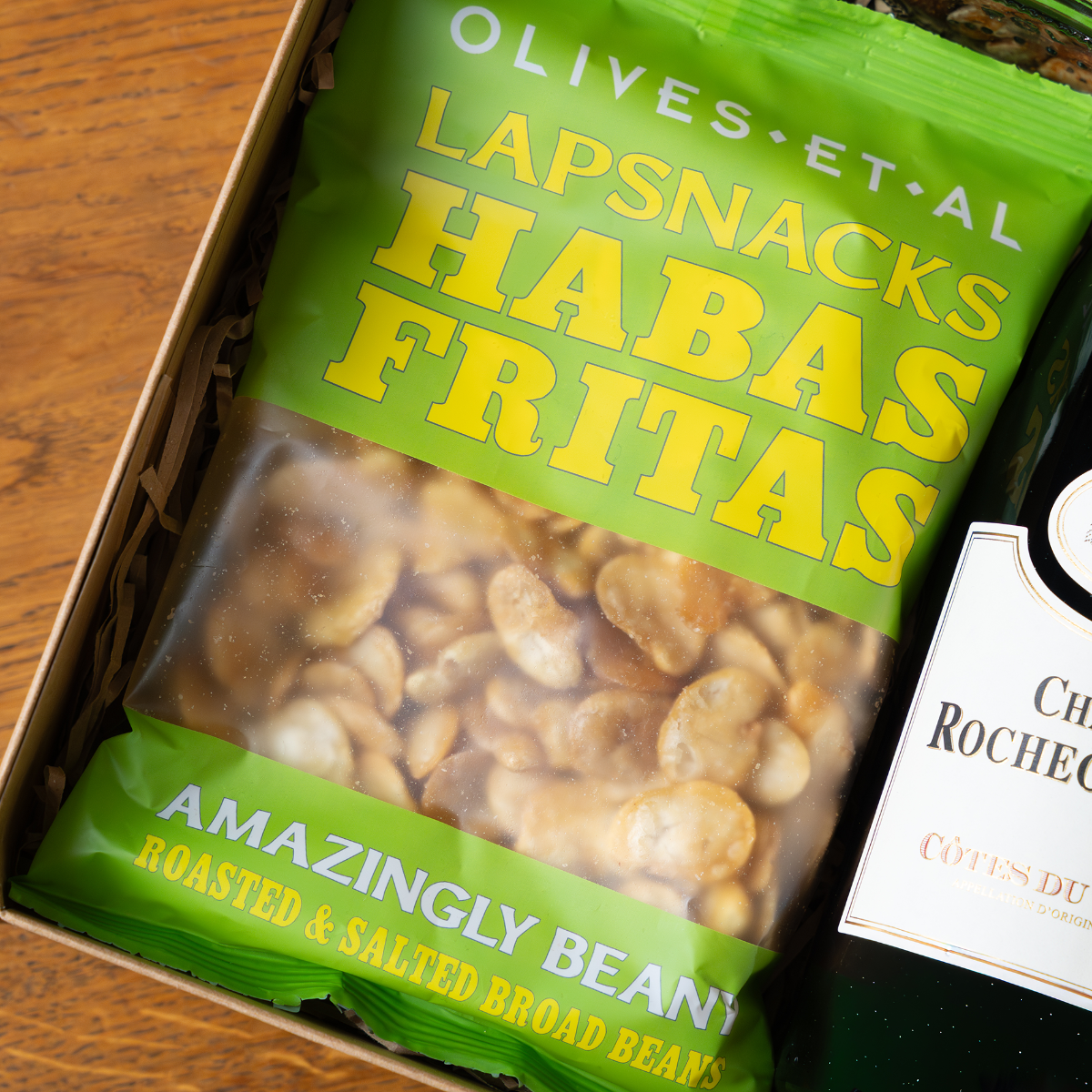 Green package of 'Lapsnacks Habas Fritas' roasted and salted broad beans on a wooden surface with a bottle of wine.