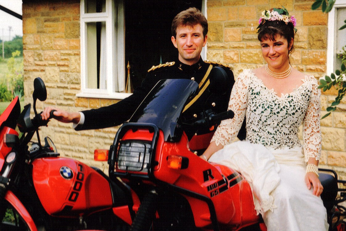 A man in a black military-style jacket with gold trim and a woman in a white lace wedding dress sit together on a red BMW motorcycle in front of a stone house.