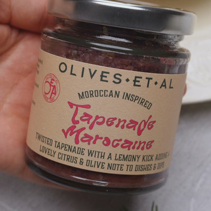 The vibrant tapenade is  generously spread onto slices of toasted bread. The scene ends with the toast being served on a wooden board in a warm, Mediterranean-style kitchen.